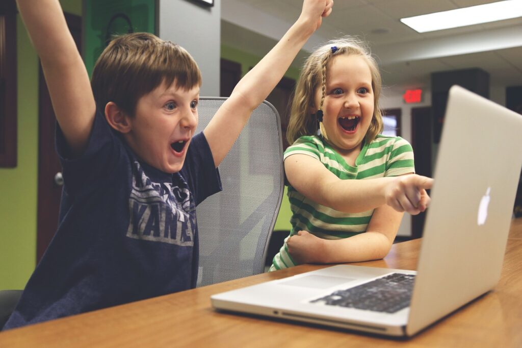 Two children smiling and pointing at a laptop.